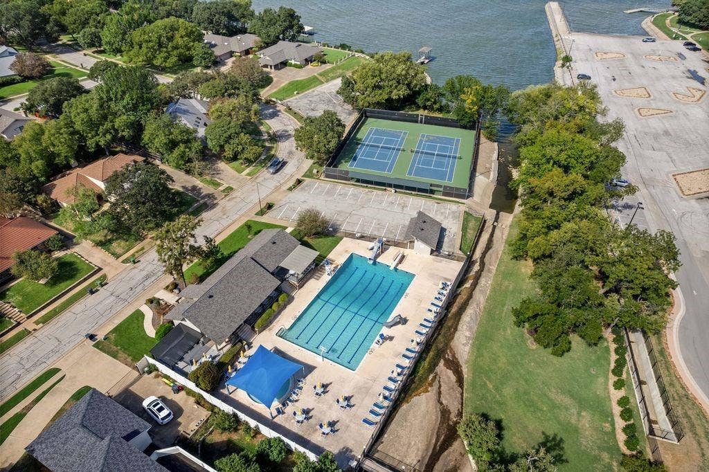 Aerial perspective of a recreational complex featuring a swimming pool, two tennis courts, shaded patio structures, and proximity to a shoreline
