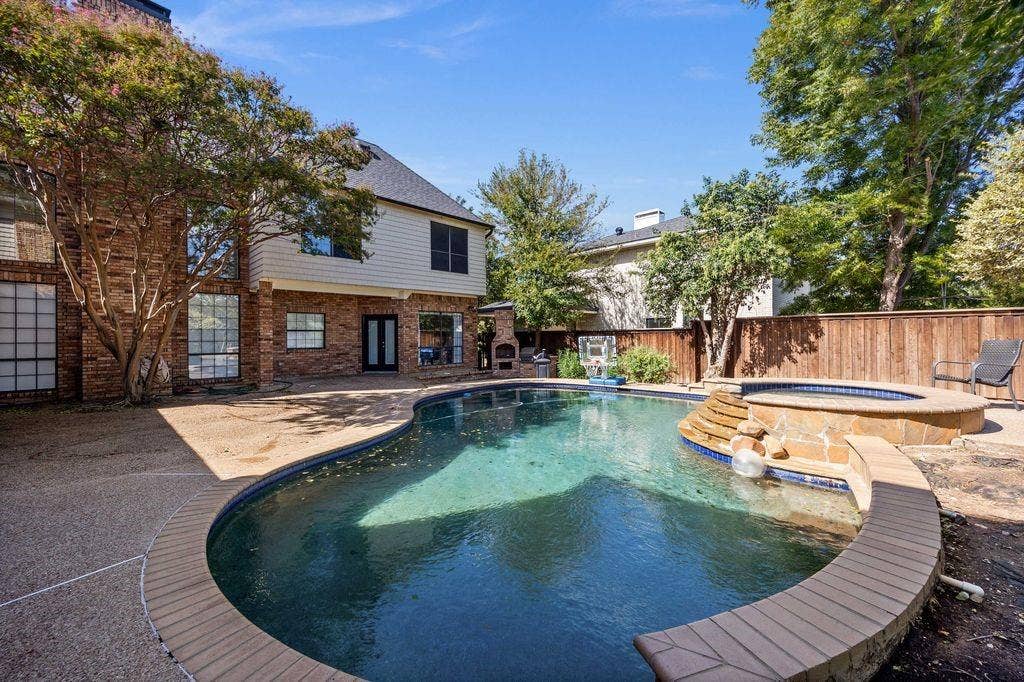 Exterior rear elevation featuring multi-textured brick and siding, custom-shaped swimming pool with integrated spa, stone-finished hardscaping, and built-in outdoor masonry fireplace