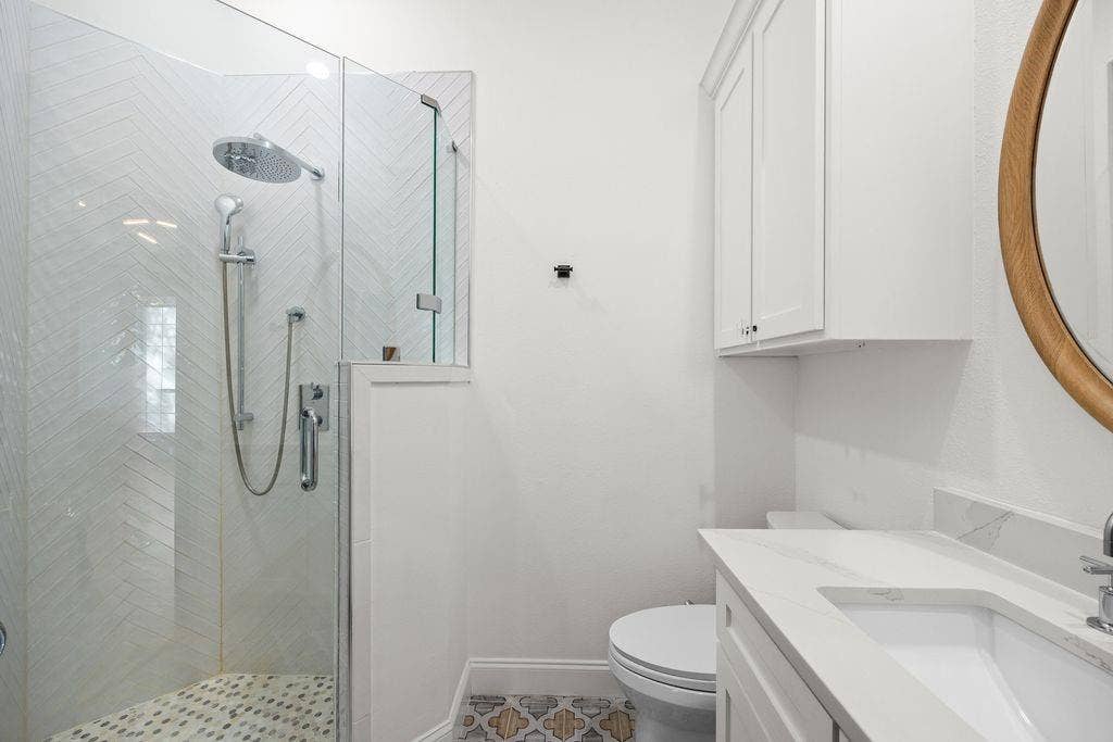 Glass-enclosed shower with chevron-patterned wall tile, polished chrome rainfall showerhead, white cabinetry with quartz vanity countertop, and decorative patterned floor tile