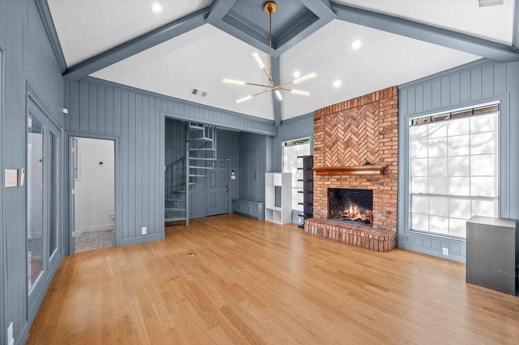 Vaulted ceiling with exposed beams, wood-finish flooring, brick fireplace with a herringbone pattern surround, metal spiral staircase, and vertical shiplap wall paneling