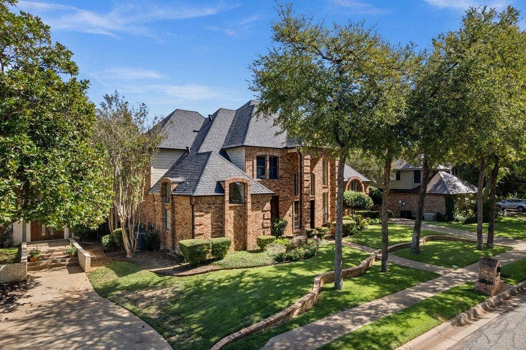 Multi-story brick exterior featuring a steep-pitched roof, stone-edged garden beds, a concrete driveway, and mature landscaping
