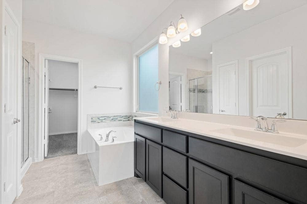 Dual-sink vanity with dark cabinetry, light countertops, soaking tub with tile backsplash, and full-width mirror