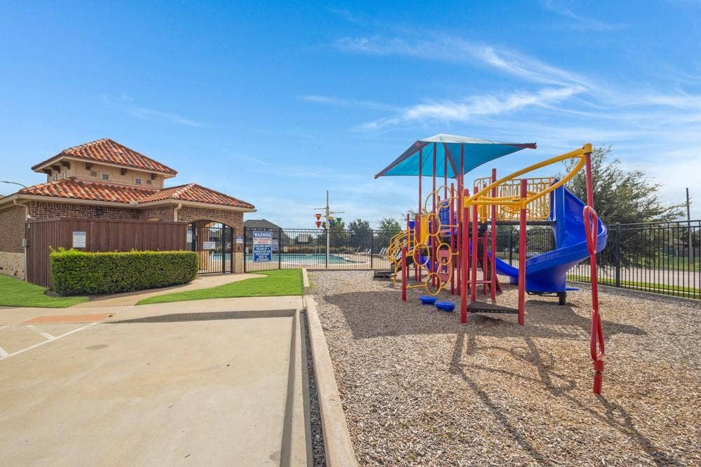 Recreational area featuring a playground with a slide, a covered structure, concrete pathways, a manicured hedge, and an outdoor swimming pool enclosed by metal fencing