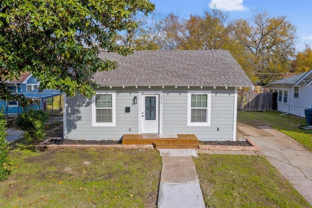 3023 Spurlock Street, Dallas, TX 75223 Single-story exterior featuring light gray horizontal siding, asphalt shingle roofing, a wood-plank front porch, and brick-edged garden beds