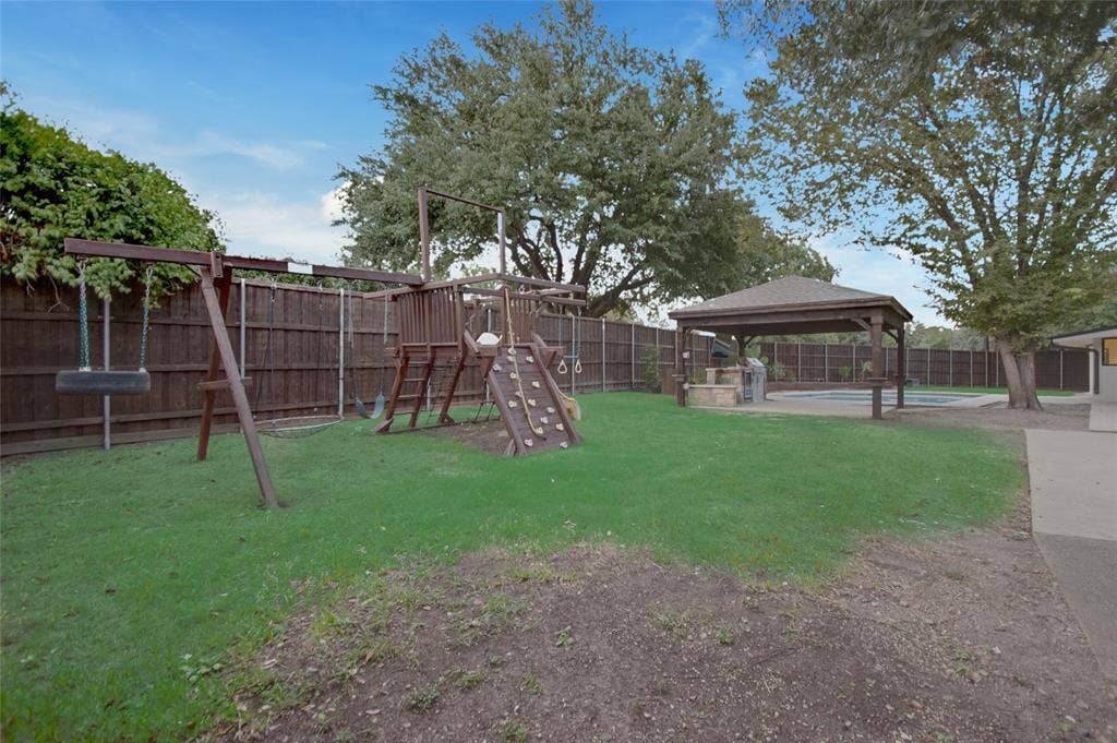 Backyard featuring a grass lawn, wooden perimeter fencing, built-in wooden play structure, and a covered pavilion with stone-base outdoor kitchen