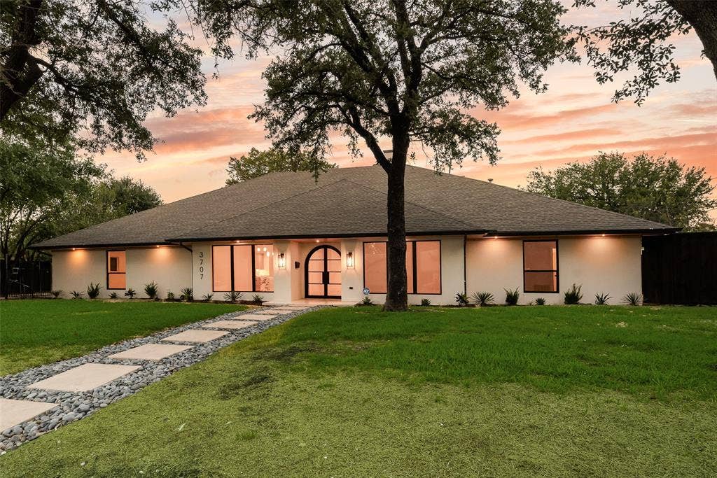 Single-story residence featuring a smooth stucco exterior, an arched iron front door with glass panels, large picture windows with black frames, and a stone-paved walkway bordered by decorative river rock