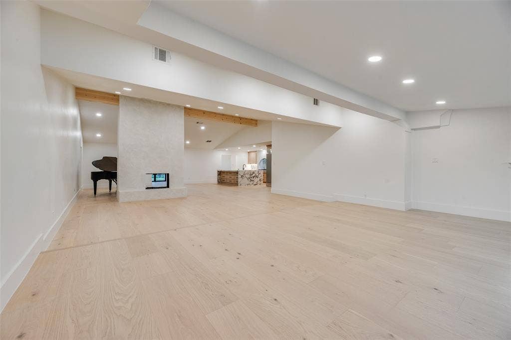 Open-concept interior featuring light wood-finish flooring, recessed lighting, exposed wood ceiling beams, and a centrally positioned masonry fireplace