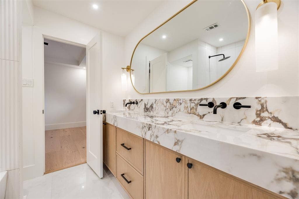 Wood-finish cabinetry with matte black hardware, stone-top dual vanity, wall-mounted matte black faucets, oval gold-rimmed mirror, and integrated cylindrical light fixtures
