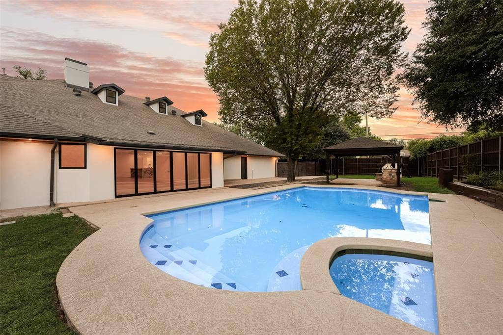Backyard featuring an in-ground swimming pool with an integrated spa, expansive concrete decking, a detached pergola, and a stucco exterior with multi-panel sliding glass doors