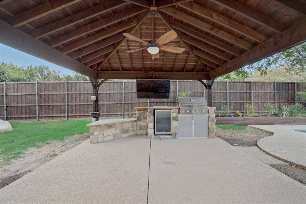 Covered outdoor kitchen pavilion featuring a timber-frame ceiling, integrated stone masonry, built-in stainless steel grill, under-counter refrigeration, and ceiling fan