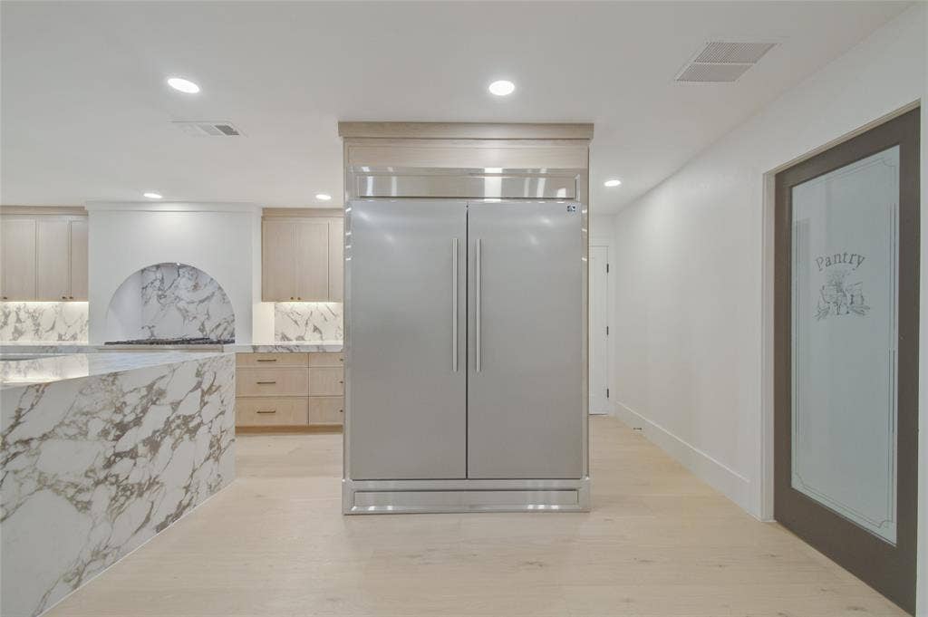 Custom cabinetry with a light wood finish, full-height built-in stainless steel refrigerator, stone countertops, integrated stone backsplash with arched detailing, and a door featuring a frosted glass panel with pantry signage