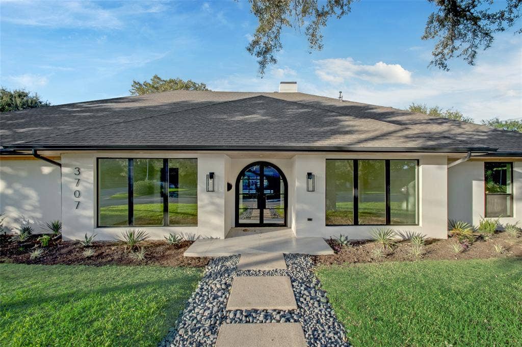 Single-story exterior featuring a light-toned stucco façade, an arched iron-framed entryway, large-pane picture windows, a multi-gabled roofline, and a concrete paver walkway bordered by decorative stone