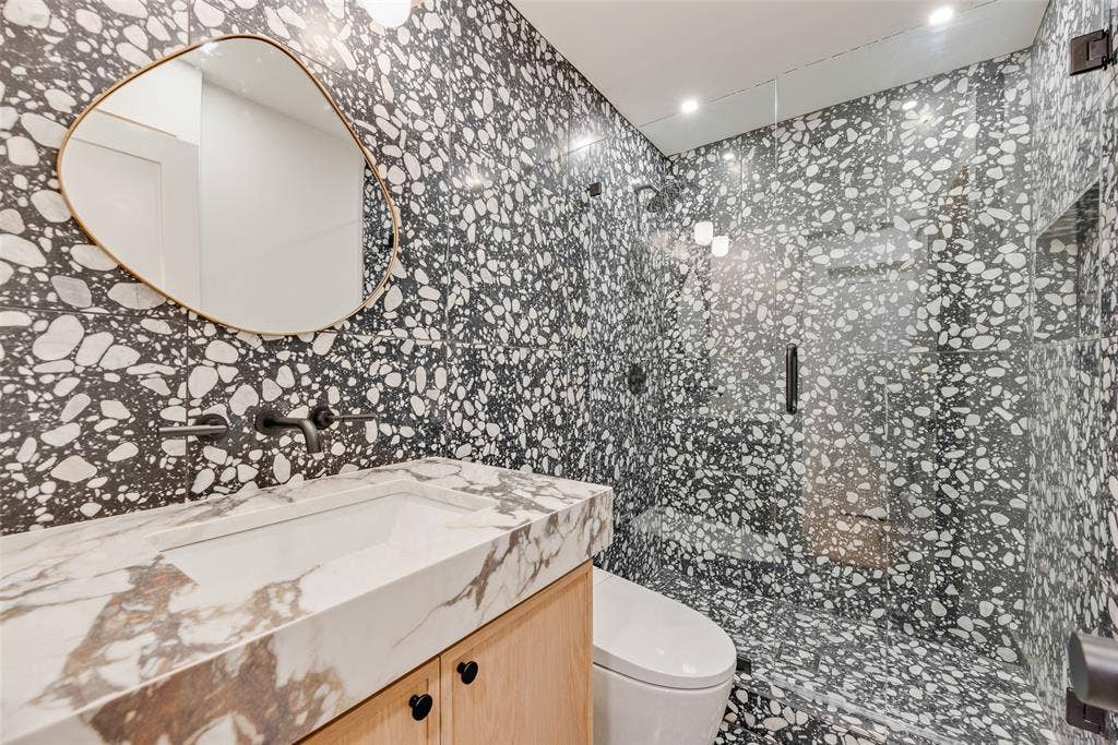 Custom vanity with stone-finish countertop, irregular-shaped wall mirror, matte black wall-mounted faucet, terrazzo-style tile wall finishes, and glass-enclosed walk-in shower