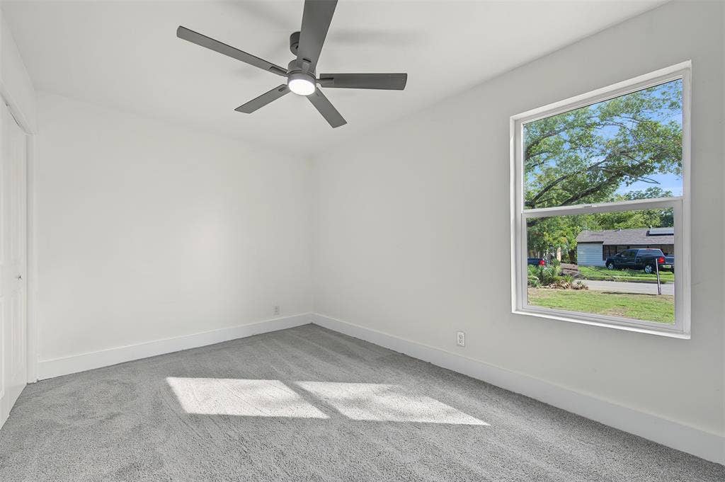 Neutral interior featuring grey carpeting, white wall finishes, a multi-blade ceiling fan with integrated lighting, and a single-hung window