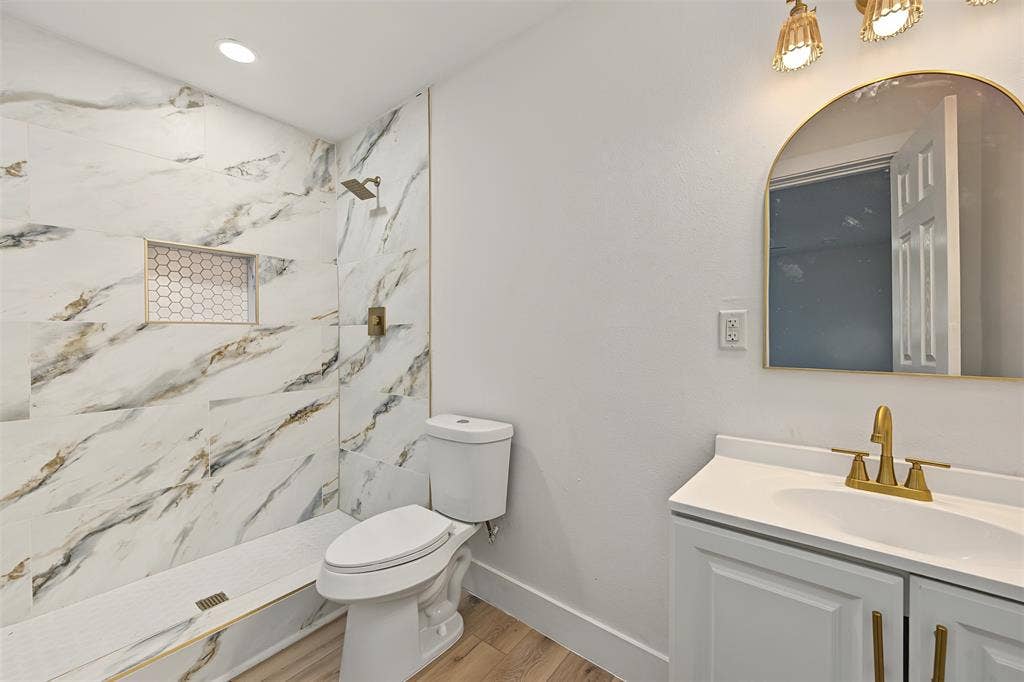 Walk-in shower featuring large-format veined tile surround, built-in geometric tile niche, and gold-tone fixtures, complemented by a vanity with a white countertop and arched gold-rimmed mirror