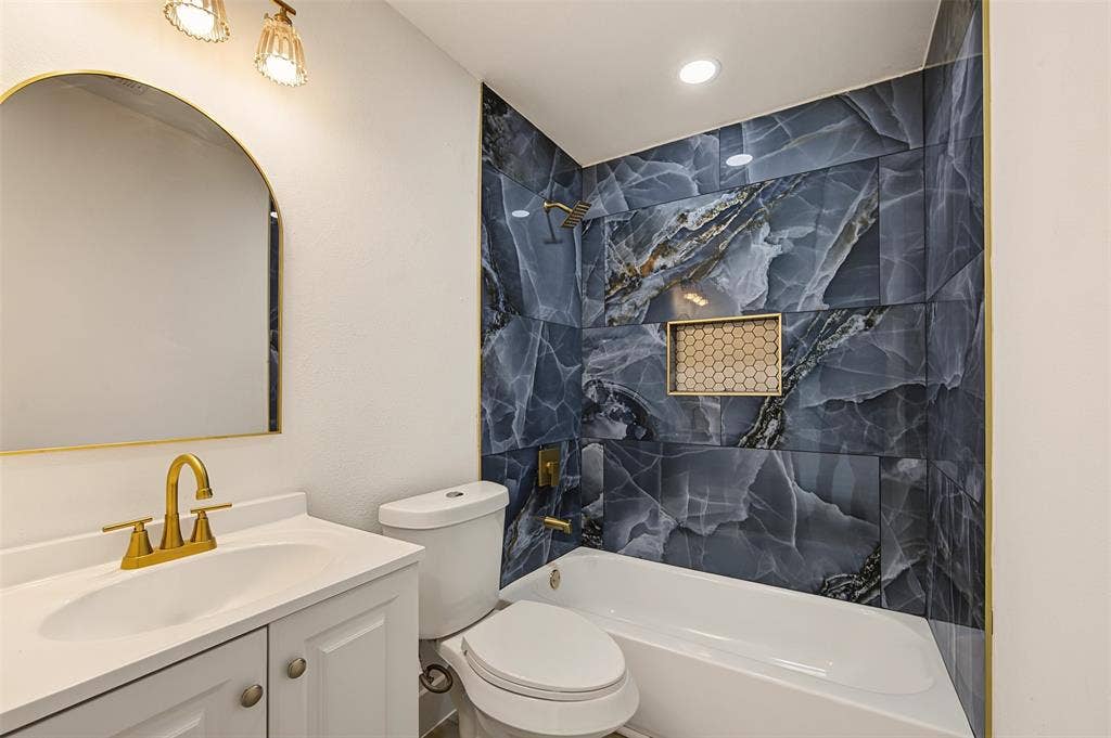 Bathroom featuring a white vanity cabinet with an integrated sink, arched mirror, brushed gold-tone hardware, and a shower-tub combination finished with large-format blue decorative tile and a hexagonal inset niche