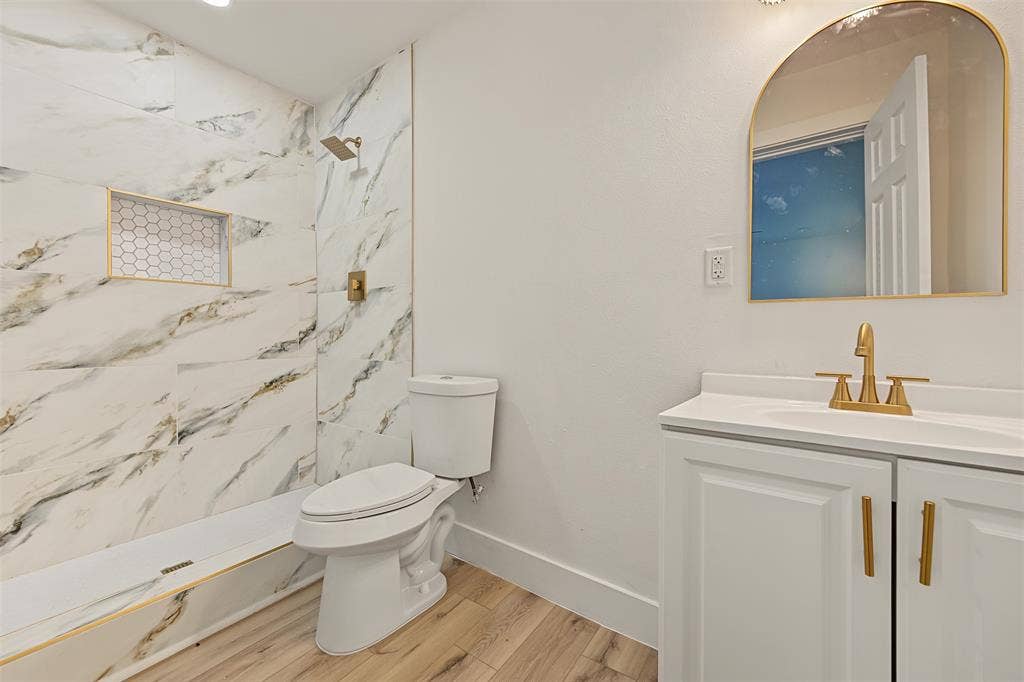 Bathroom featuring floor-to-ceiling veined tile, wood-finish flooring, a vanity with gold-toned fixtures, a built-in shower niche with hexagon tile, and an arched mirror