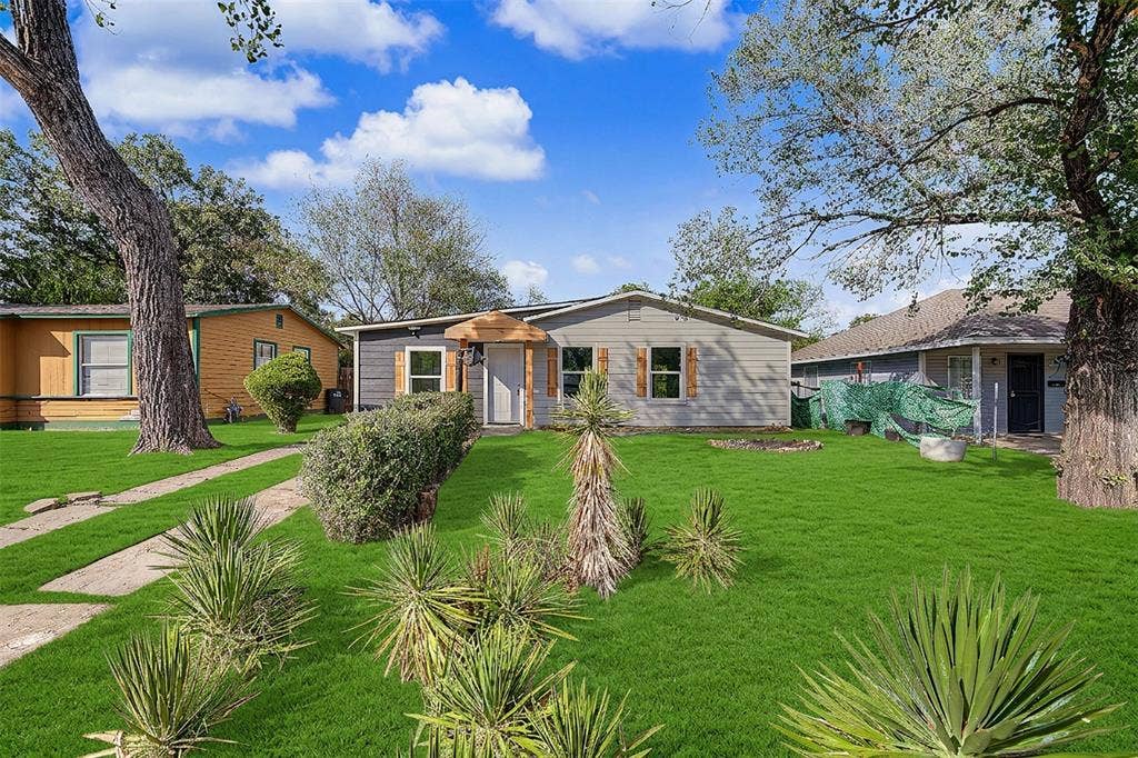 Single-story exterior featuring gray horizontal siding, wood-toned shutter accents, a covered entryway with wood architectural detailing, and mature landscaping