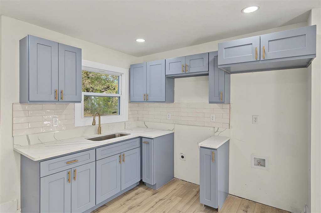 Kitchen featuring blue shaker-style cabinetry, light-toned stone-finish countertops, subway tile backsplash, brass-finish hardware, and wood-finish flooring