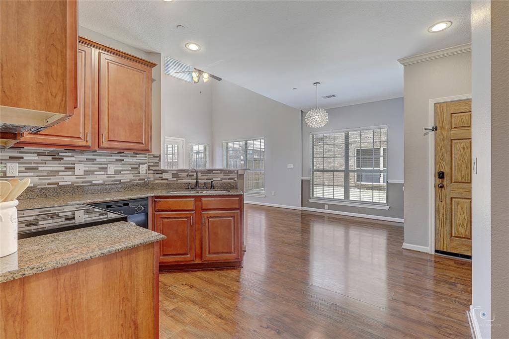 Kitchen featuring stained wood cabinetry, stone-finish countertops, and a multi-toned tile backsplash, complemented by wood-finish flooring, vaulted ceilings, and a decorative pendant light fixture