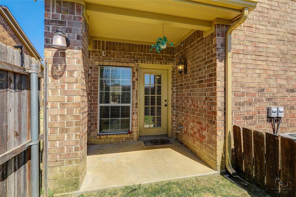 Covered brick patio featuring a glass-paneled entry door, single-hung window with grid inserts, and wall-mounted exterior lighting fixtures