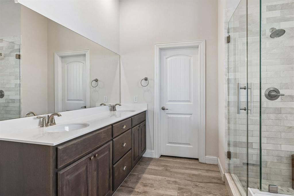 284 Goliad Trail, Forney, TX 75126 Dual-sink vanity with a solid-surface countertop, full-width wall mirror, wood-finish flooring, and a glass-enclosed shower featuring stone-style wall tile