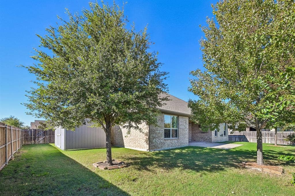 284 Goliad Trail, Forney, TX 75126 Rear exterior featuring brick siding, a concrete patio, mature trees, a manicured lawn, and wood-panel fencing