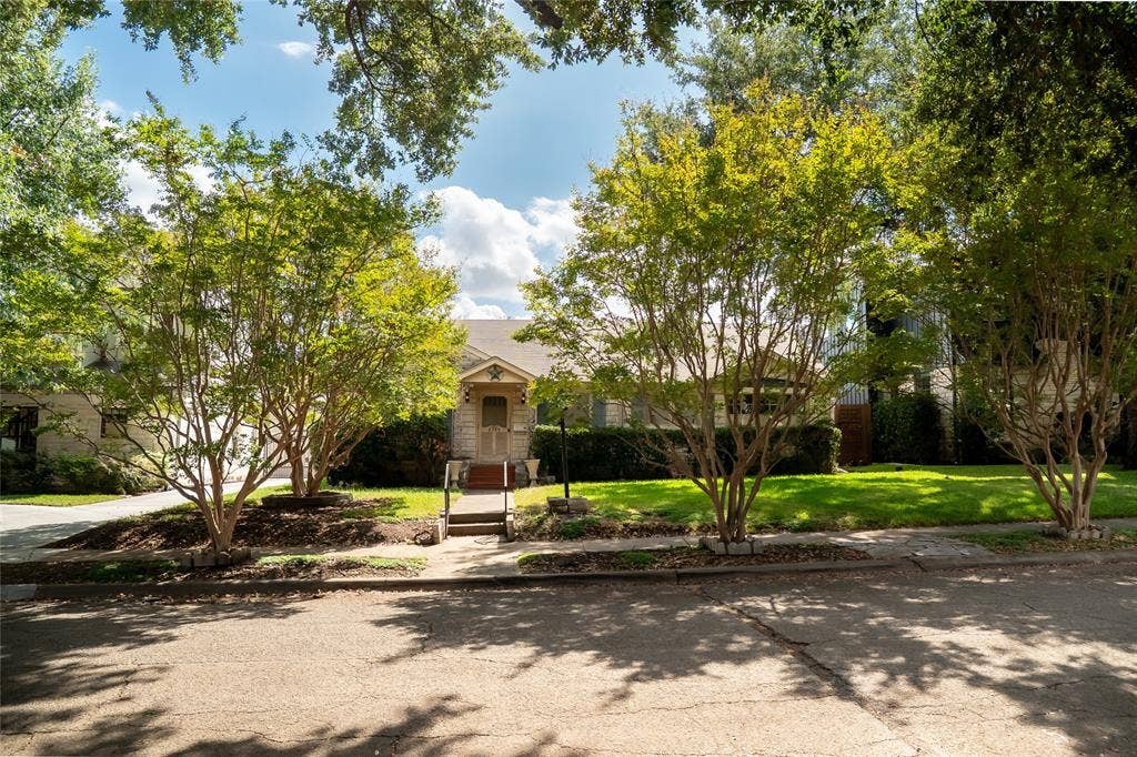 Single-story stone exterior featuring a gabled roofline, concrete walkway, mature specimen trees, and established landscape beds