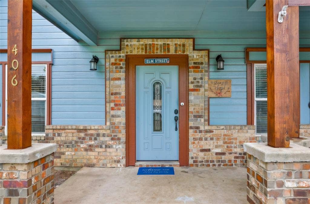 Covered front porch featuring a multi-toned brick facade, wood-finish support columns, horizontal siding, and a central entry door with a decorative glass insert