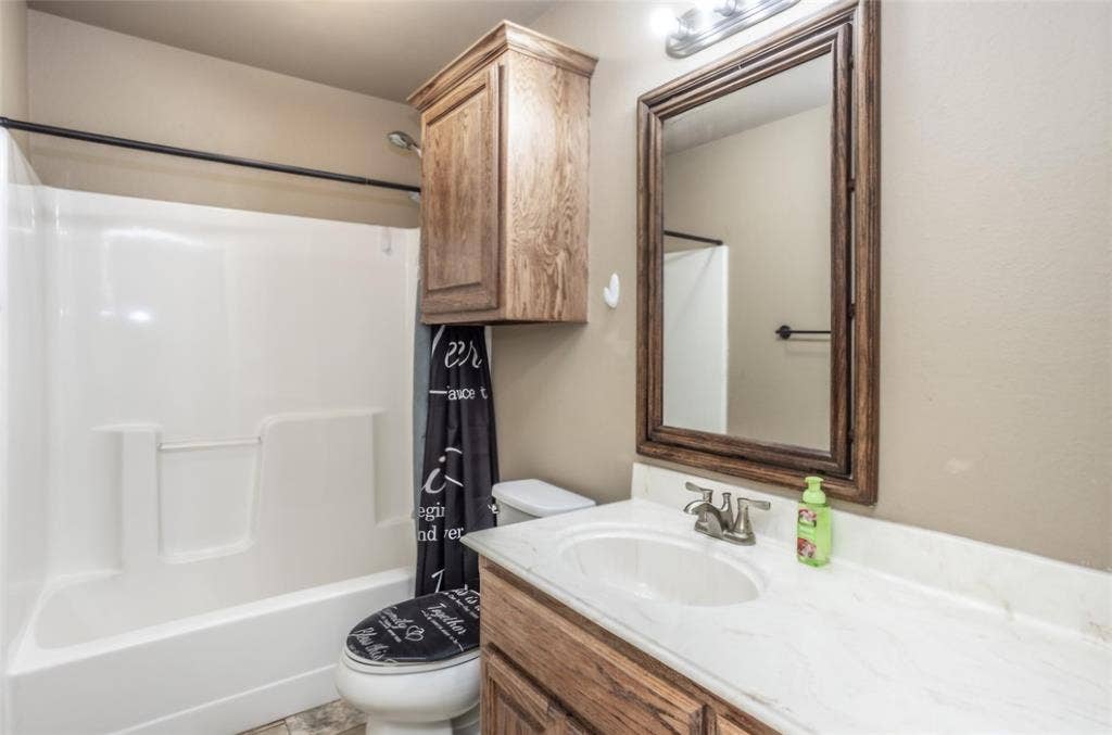 Bathroom featuring a white molded tub-shower surround, wood-finish vanity with a stone-composite countertop, matching wall-mounted cabinet, and wood-framed vanity mirror