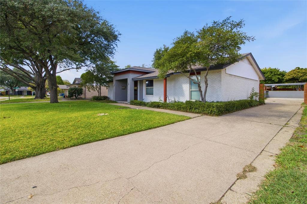 This perspective showcases the home's attractive white brick exterior, clean architectural lines, and mature shade trees that complement its contemporary styling. The extended driveway runs alongside the home, providing convenient access to the attached two-car garage and gated rear parking area. Established landscaping and the generous front lawn add to the property's inviting curb appeal.