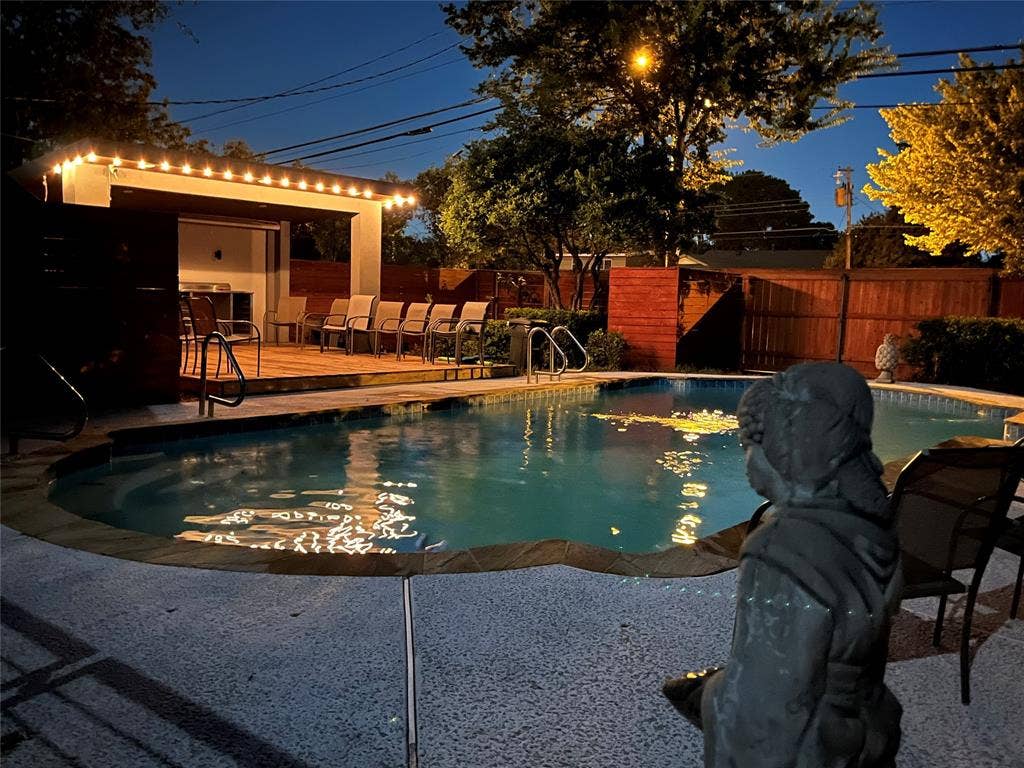 As evening settles in, the backyard transforms into an inviting poolside retreat. Warm string lighting outlines the spacious covered cabana, casting beautiful reflections across the swimming pool. With comfortable seating, mature trees, and privacy fencing surrounding the space, this outdoor setting offers a welcoming atmosphere for relaxing after sunset or gathering with friends under the evening sky.