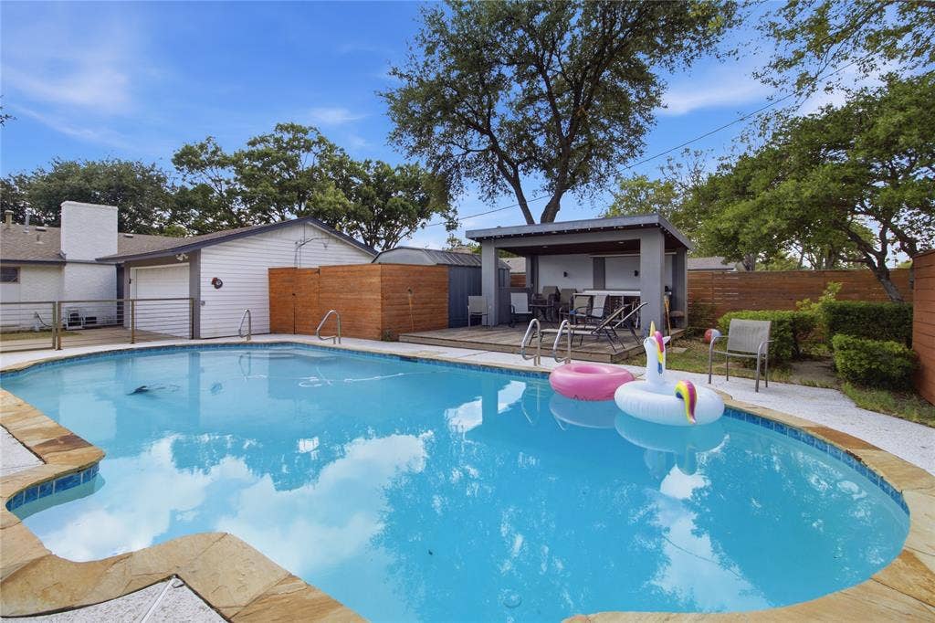 This perspective highlights the relationship between the sparkling swimming pool, spacious covered cabana, and surrounding deck areas. The cabana provides a comfortable shaded space just steps from the water, while the surrounding deck offers room for poolside seating and relaxation. Mature trees and wood privacy fencing complement the setting, creating an inviting backyard retreat with several distinct outdoor spaces to enjoy.
