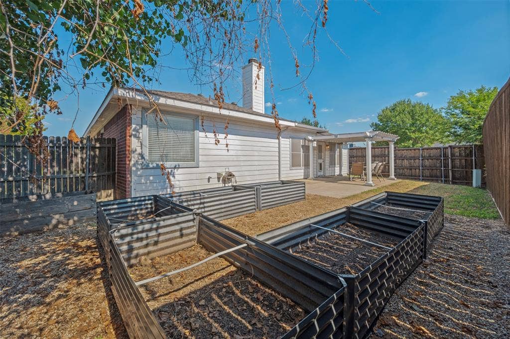Backyard featuring elevated metal garden beds, a concrete patio, a covered pergola with columns, a masonry chimney, and perimeter wood fencing