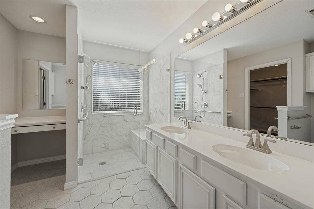 Dual-sink vanity with white cabinetry and solid-surface countertops, walk-in shower with integrated bench and wall-mounted spray fixture, hexagon-patterned tile flooring, and large-format wall tiling