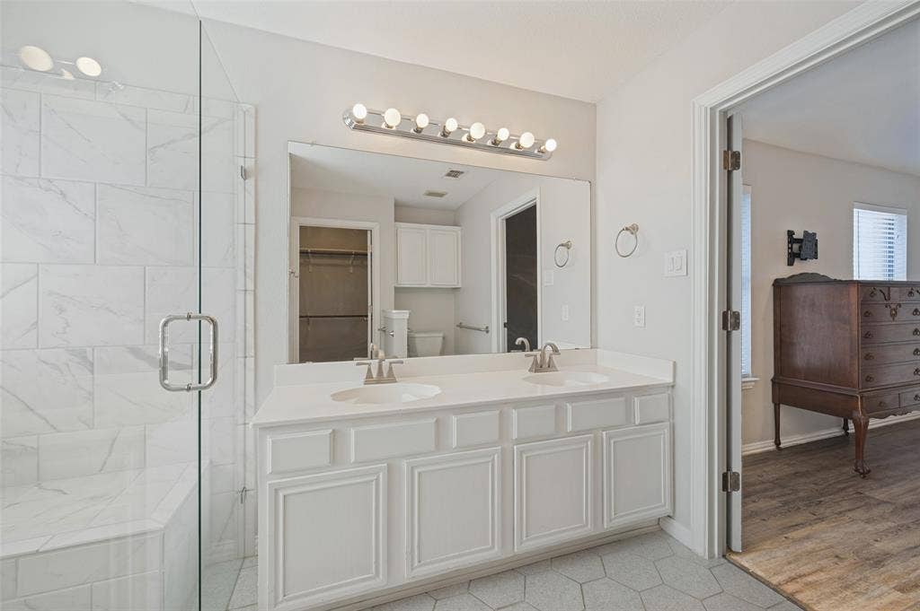 Dual-sink vanity with white cabinetry and countertop, large wall-mounted mirror, multi-bulb vanity lighting, and walk-in shower with stone-patterned tile surround