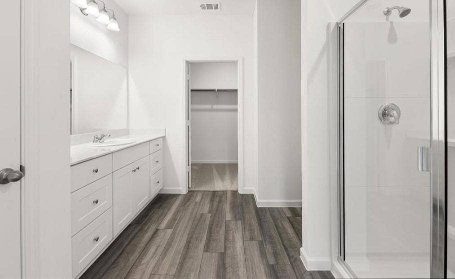 Bathroom featuring a white shaker-style vanity with stone-look countertop, wood-finish flooring, glass-enclosed shower, and built-in walk-in closet shelving