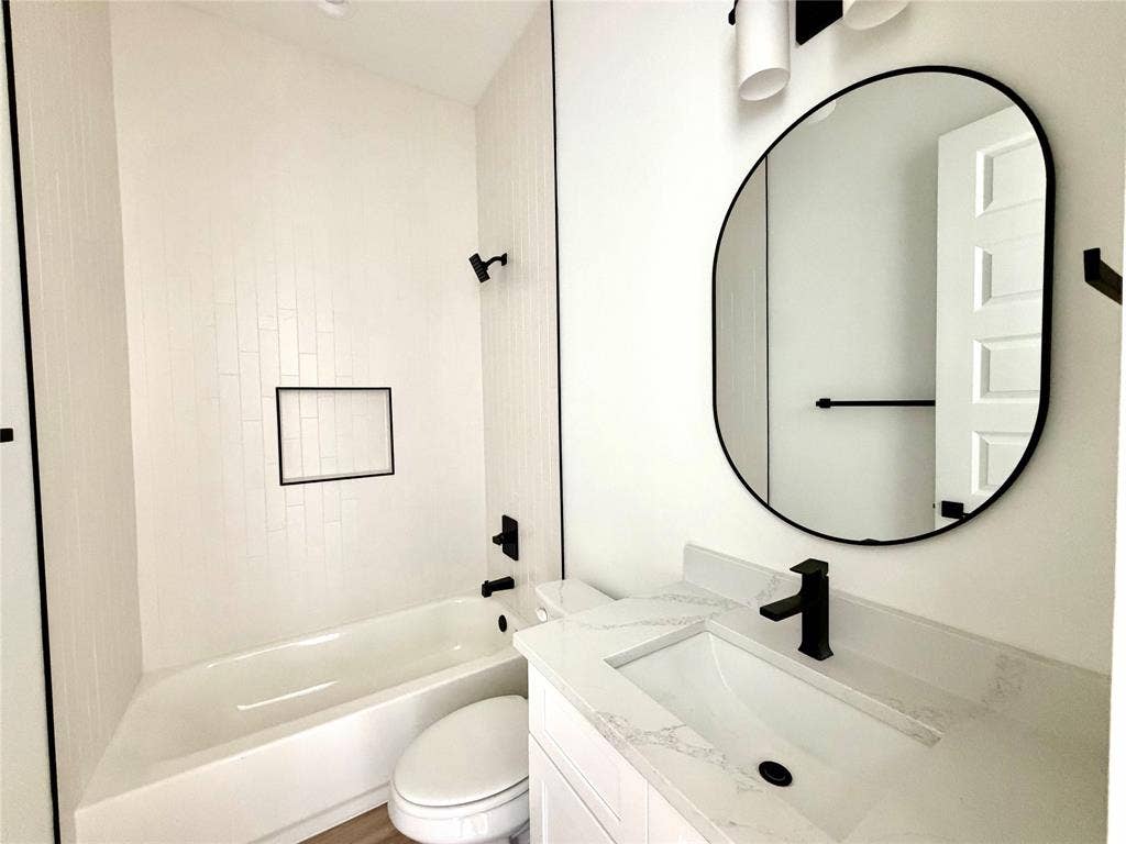 8902 Lowell Street, Rowlett, TX 75089 Bathroom featuring a vanity with a white countertop and rectangular undermount sink, matte black faucet, wall-mounted oval mirror, built-in tub with vertical white wall tile, and recessed shower niche