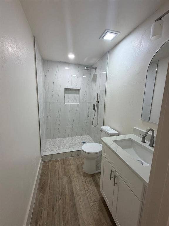 Bathroom featuring wood-finish flooring, white vanity with stone-style countertop, arched mirror, and walk-in shower with stone-look wall tiles, built-in recessed shelf, and pebble-style tile shower floor