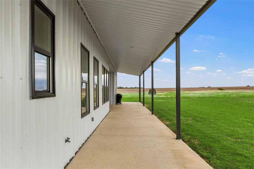 Covered concrete patio featuring a metal exterior wall, dark-framed windows, and metal structural supports overlooking an expansive open field