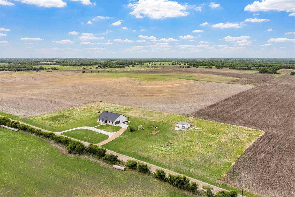 Aerial perspective of a sprawling rural property featuring a single-story residence, a detached secondary structure, a concrete driveway, and expansive acreage