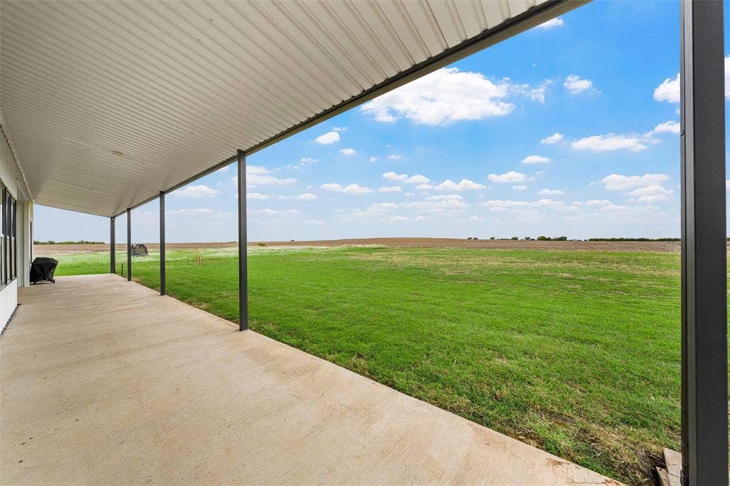 Covered concrete patio featuring a corrugated metal ceiling and metal support columns, overlooking an expansive open lawn