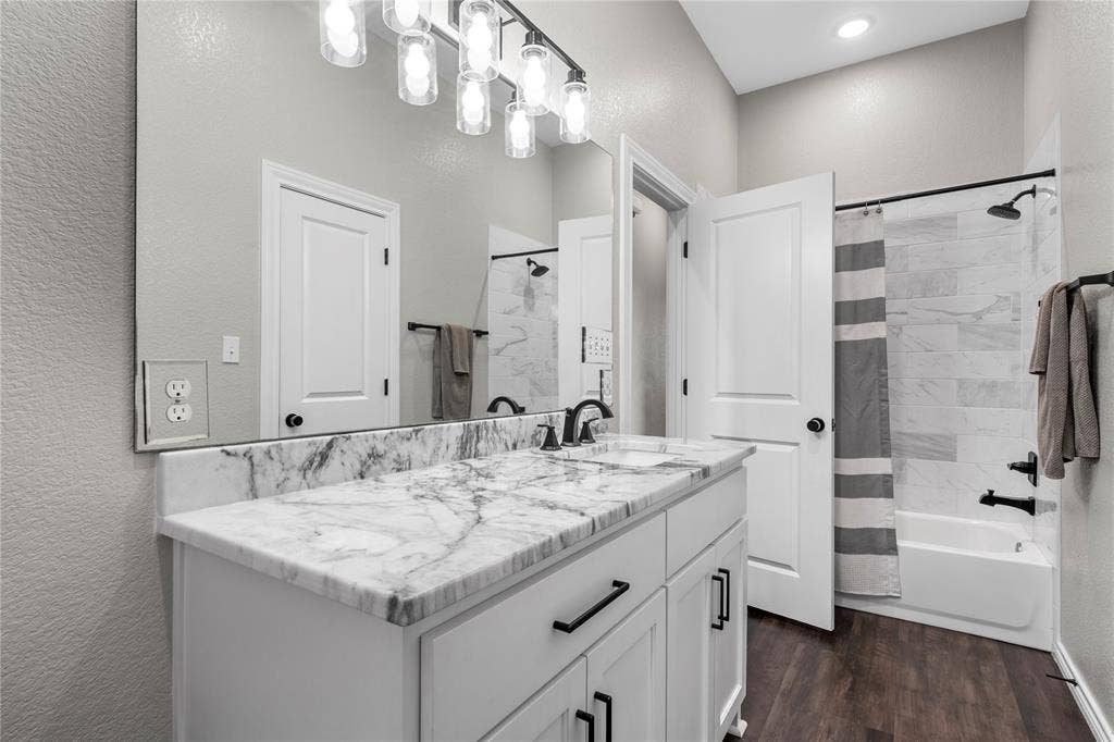 Bathroom featuring a stone-top vanity with undermount sink, matte black fixtures, full-width wall mirror, wood-finish flooring, and a tiled tub-shower combination