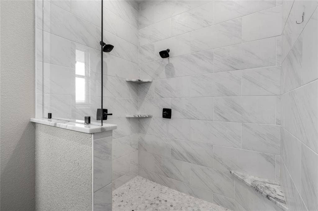 Walk-in shower featuring floor-to-ceiling rectified-edge tile, a frameless glass partition, matte black rainfall showerhead, dual built-in corner shelving, and a pebble-stone basin floor