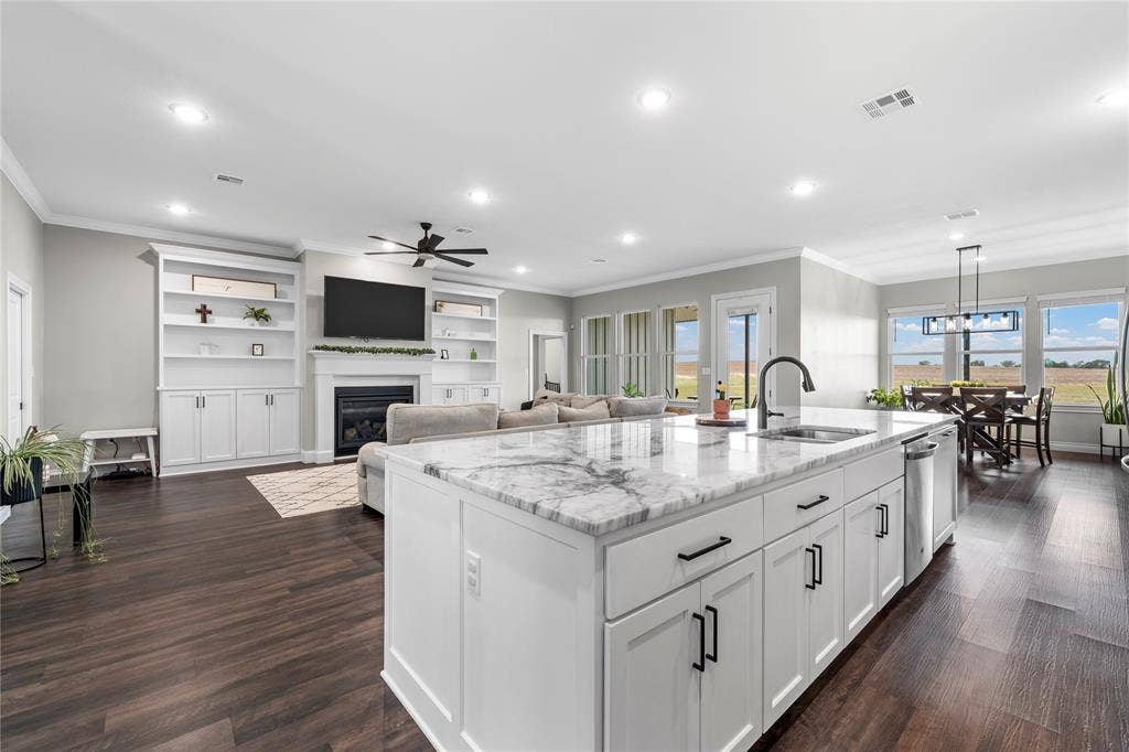 Open-concept interior featuring an oversized kitchen island with a stone countertop, white cabinetry with matte black hardware, recessed lighting, built-in shelving flanking a fireplace, and wood-finish flooring