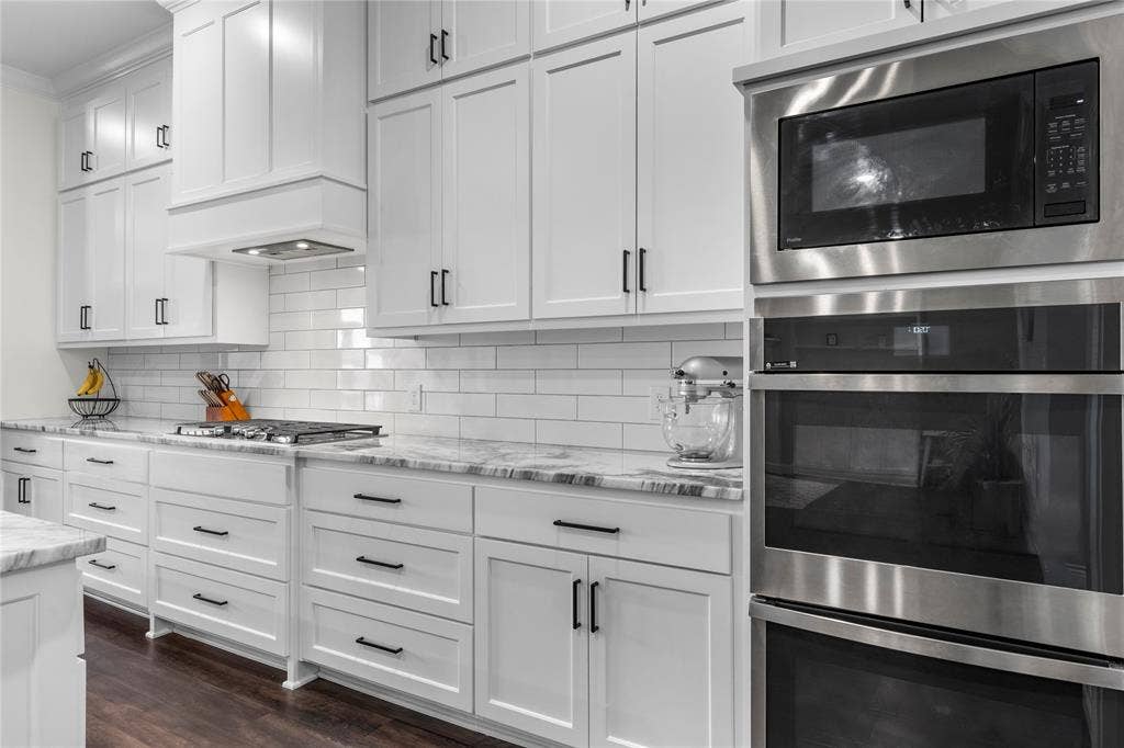 Kitchen featuring ceiling-height white shaker cabinetry, stone countertops, a subway tile backsplash, built-in stainless steel double ovens, and wood-finish flooring