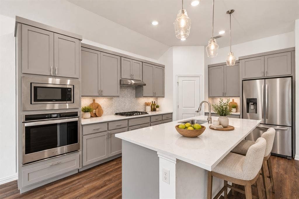 2305 Switchgrass Street, Melissa, TX 75454 Kitchen featuring grey shaker-style cabinetry, a quartz-topped center island with a stainless steel undermount sink, herringbone-patterned tile backsplash, stainless steel built-in double oven and microwave, and wood-finish flooring. Virtual photo