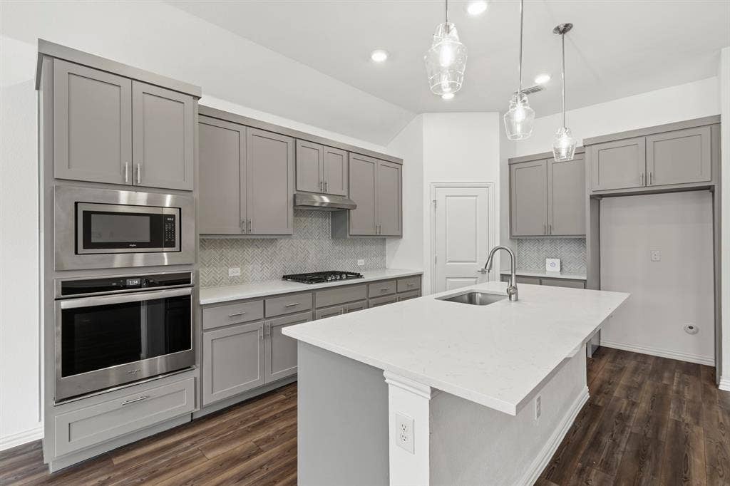 2305 Switchgrass Street, Melissa, TX 75454 Kitchen featuring grey shaker-style cabinetry, a central island with a quartz countertop, stainless steel built-in appliances, herringbone tile backsplash, and wood-finish flooring