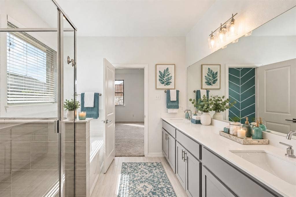 2305 Switchgrass Street, Melissa, TX 75454 Bathroom featuring a dual-sink vanity with stone countertop, frameless glass-enclosed shower, soaking tub, and wall-to-wall mirror with four-light fixture