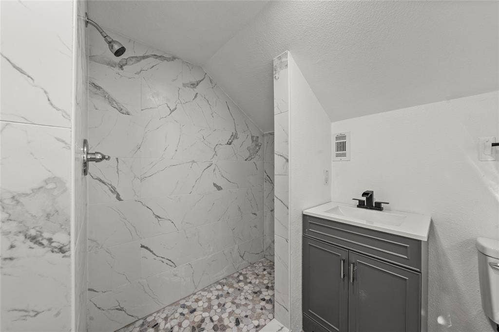 Walk-in shower with stone-patterned wall tile and pebble flooring, featuring a dark gray vanity, integrated sink, and matte black hardware